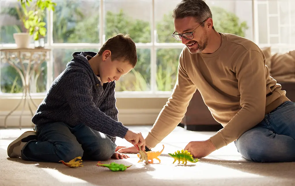 A tutor and child play with toy dinosaurs indoors.