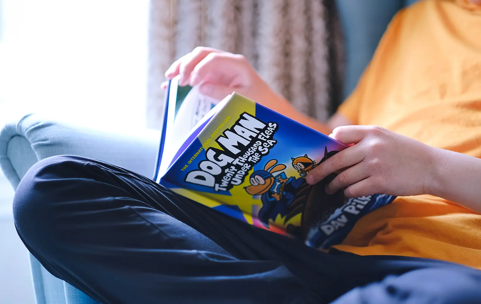 Child reading "Dog Man: Twenty Thousand Fleas Under the Sea" on a cozy couch.