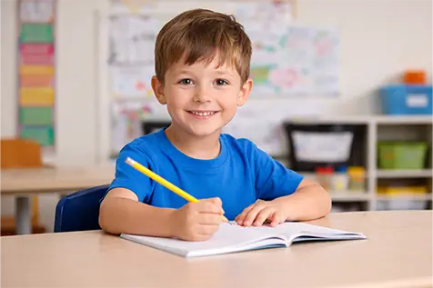 Young boy smiling while writing in a notebook at a desk.